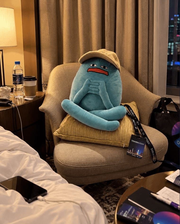 Blob wearing a beige cap, sitting cross-legged on a hotel armchair next to a conference lanyard