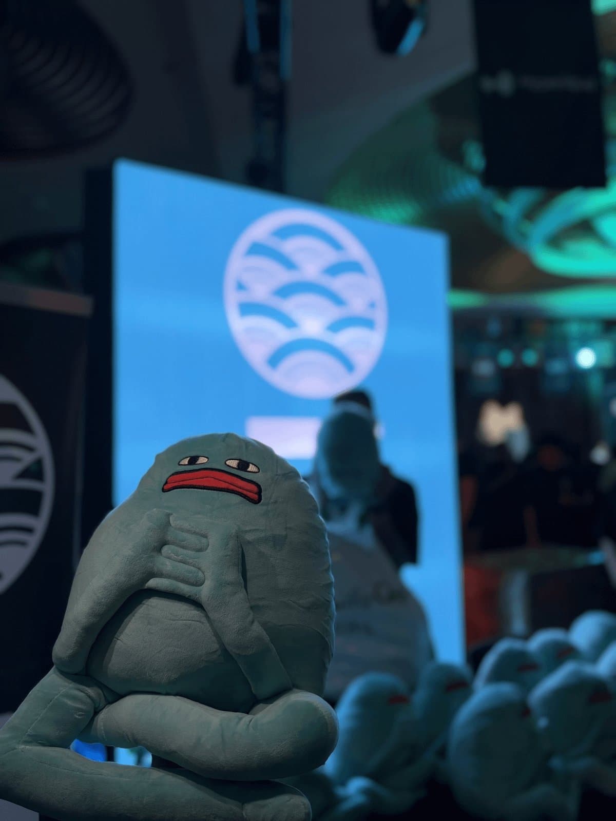 Blob sitting in front of a stage with a big blue screen, a pile of other Blobs in the background