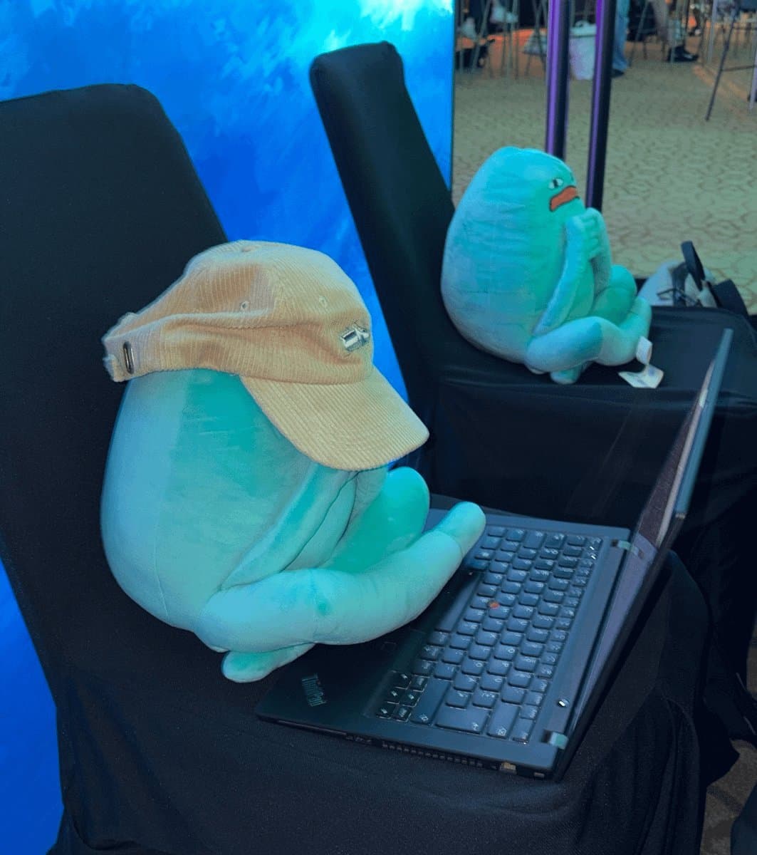 Blob in a beige cap sitting at a laptop on a chair, with another Blob watching from the next seat