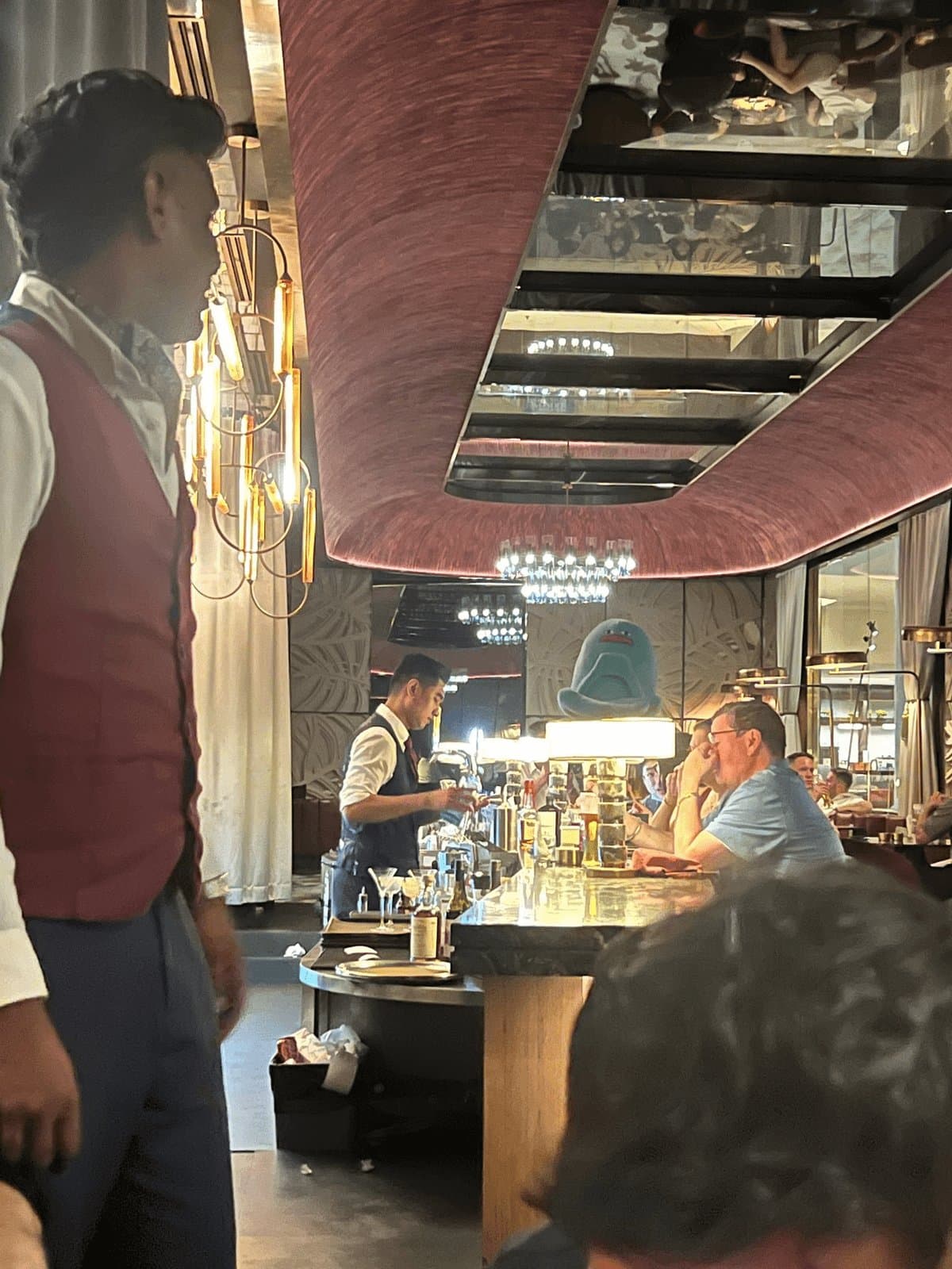 Blob perched on top of a lamp at a busy cocktail bar while a bartender mixes drinks
