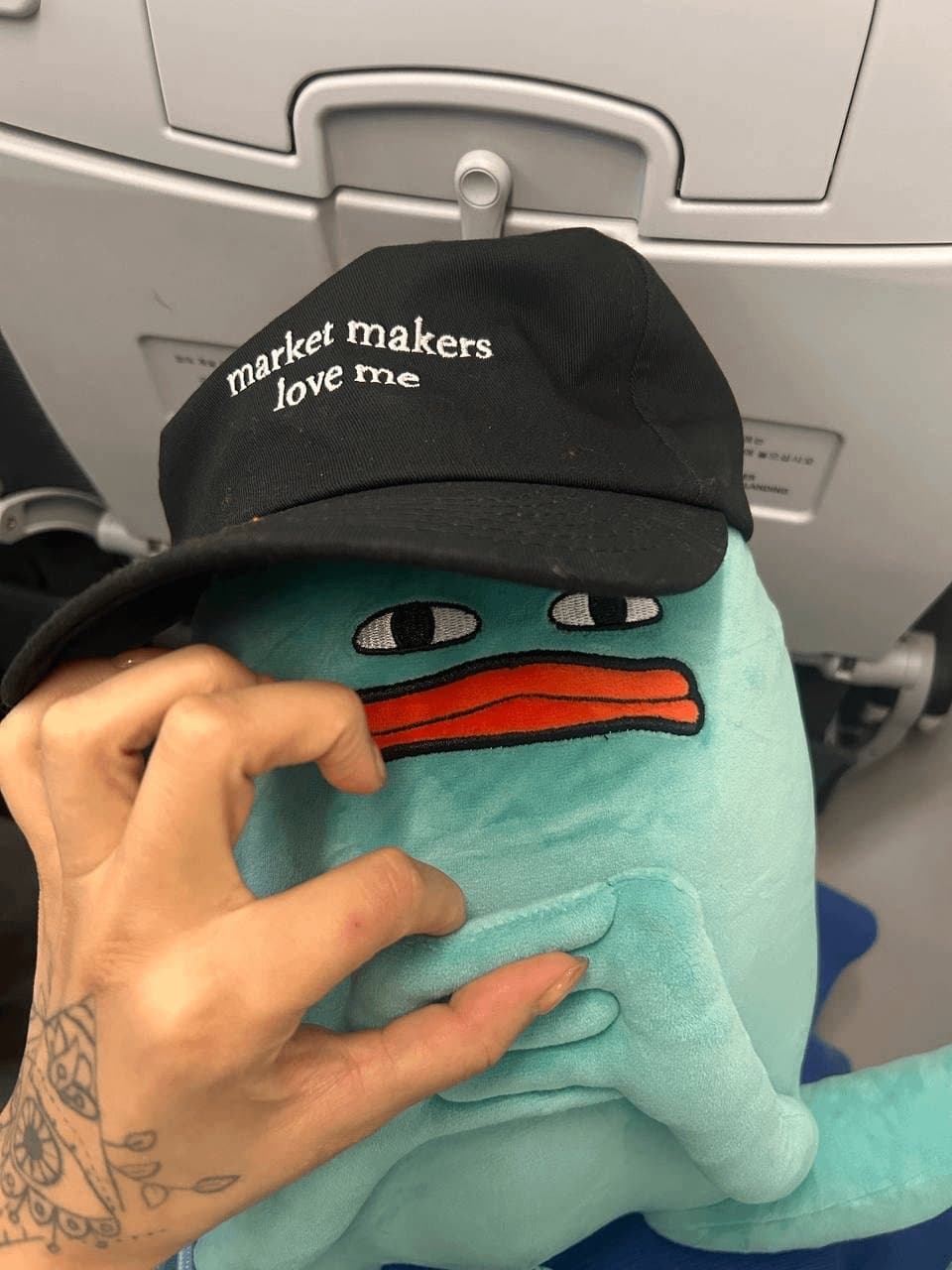 Blob on an airplane seat wearing a black cap that reads 'market makers love me'